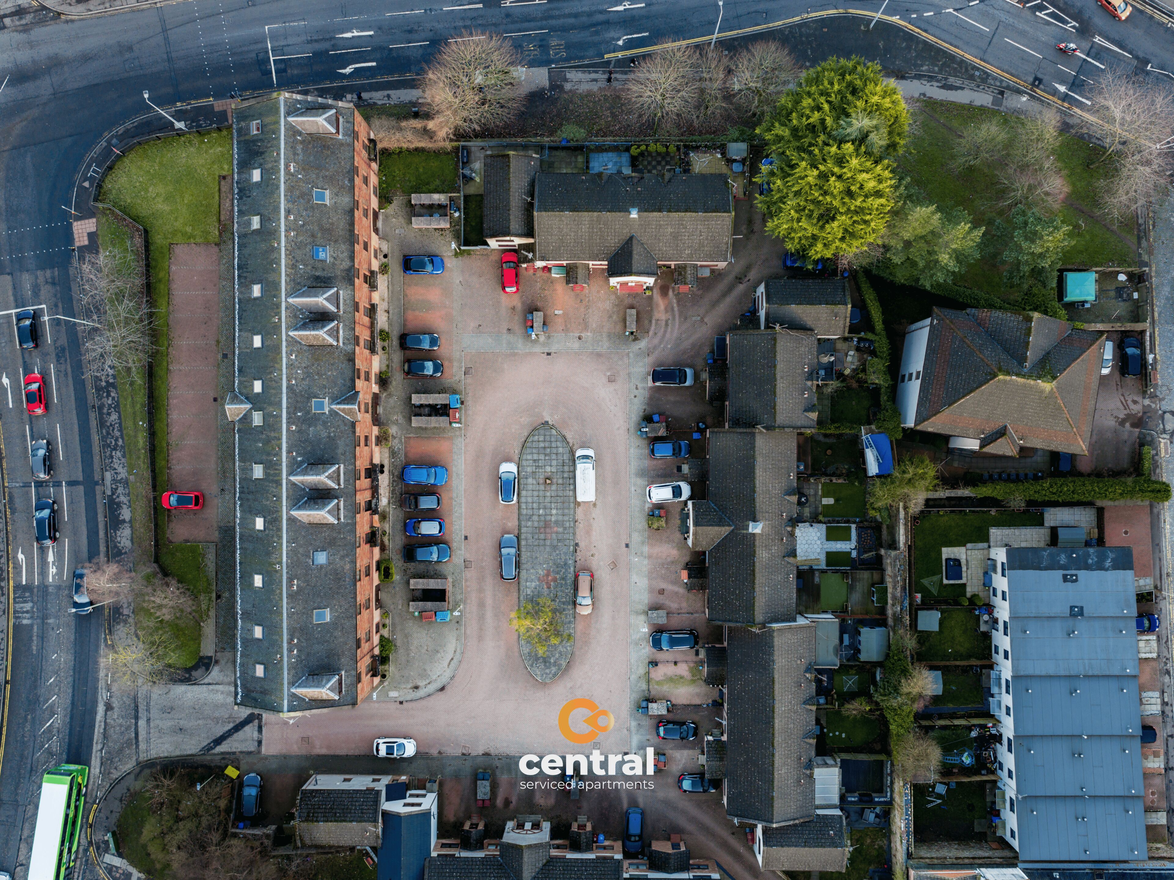 Vue aérienne d’un ensemble d’appartements avec services, comprenant un parking central, plusieurs bâtiments et une route où circulent des véhicules.