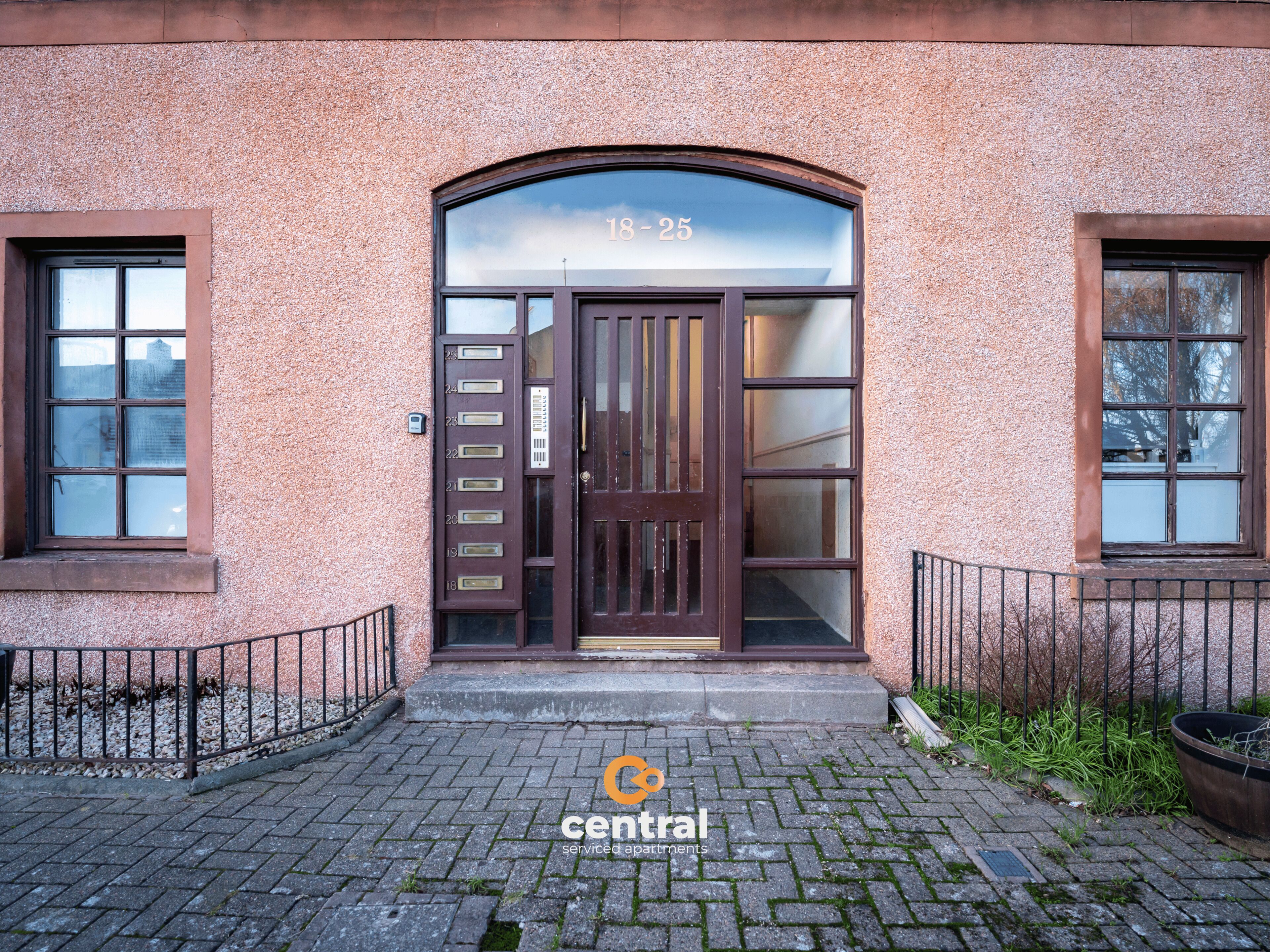 A building entrance with a brown door and numbered mail slots, a brick pavement, and a metal fence.