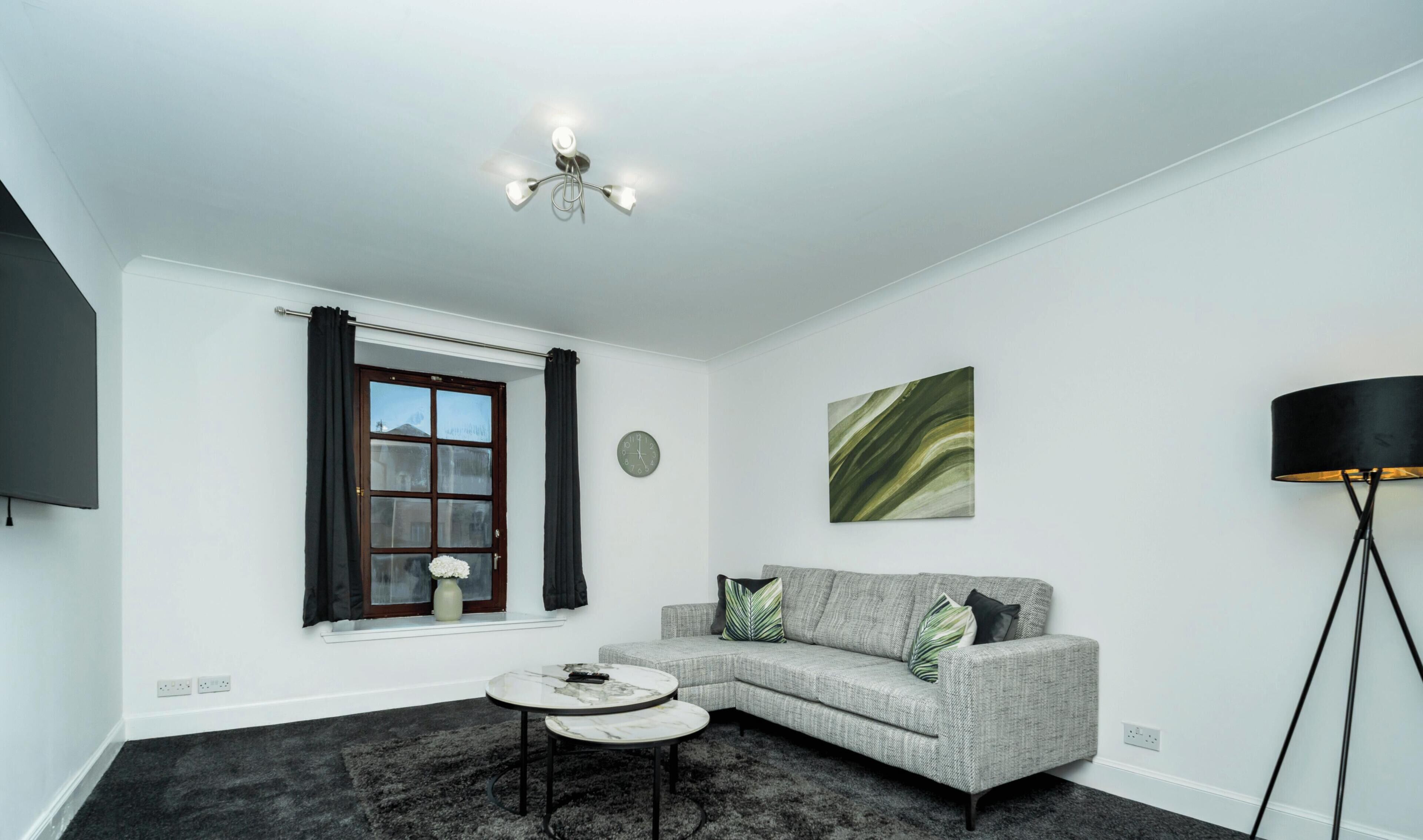 A modern living room with a grey sofa, a round coffee table, a floor lamp, a window with curtains, and a wall clock.