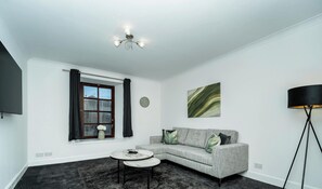 Una sala de estar moderna con un sofá gris, una mesa de centro redonda, una lámpara de pie, una ventana con cortinas y un reloj de pared.