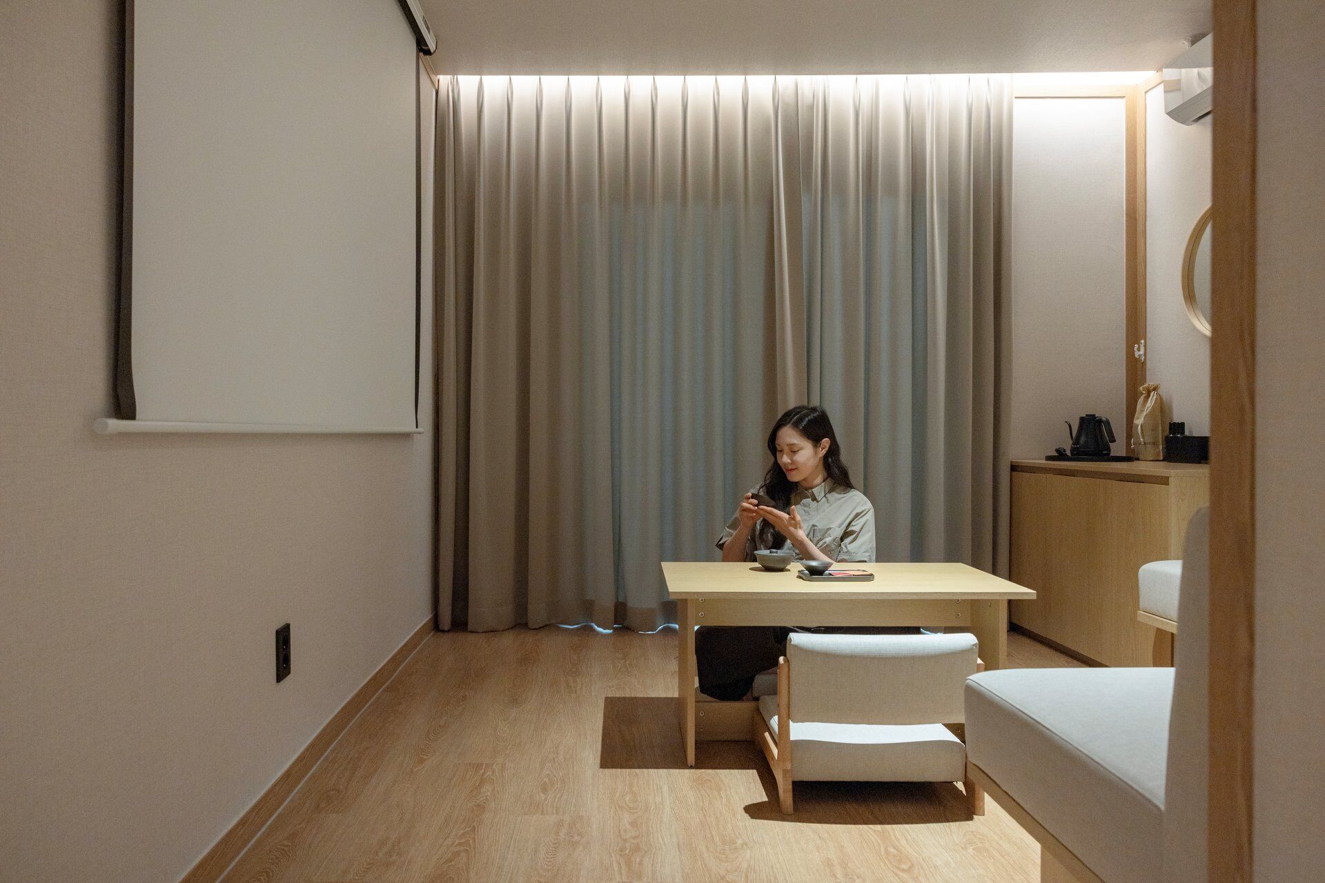 A room with a wooden desk, a chair, a bed, a small table with a kettle, and a mirror.