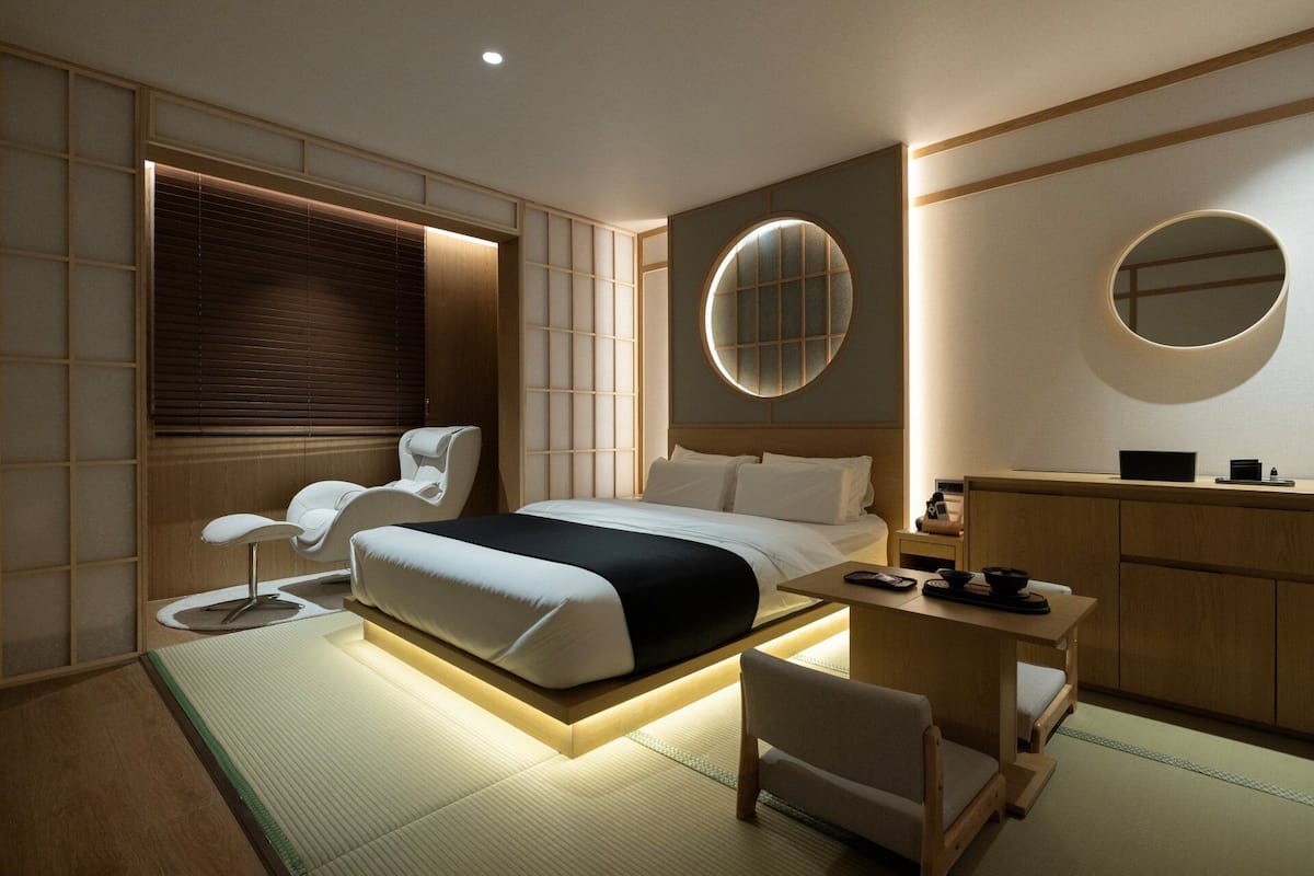 A modern hotel room with a bed, a massage chair, a desk, and a vanity with a mirror.