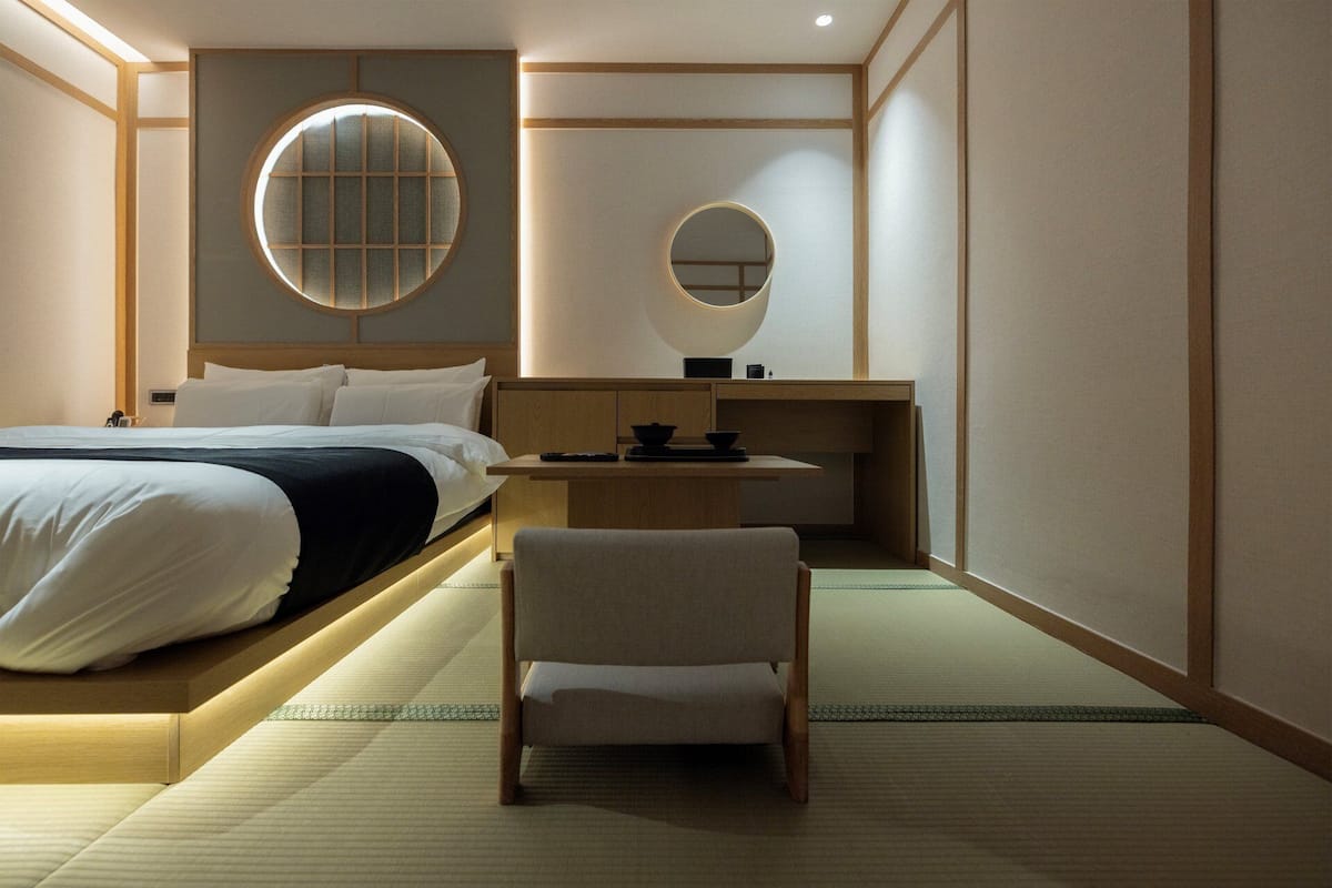 A hotel room with a bed, a round mirror, a small table with a bowl, and a chair.
