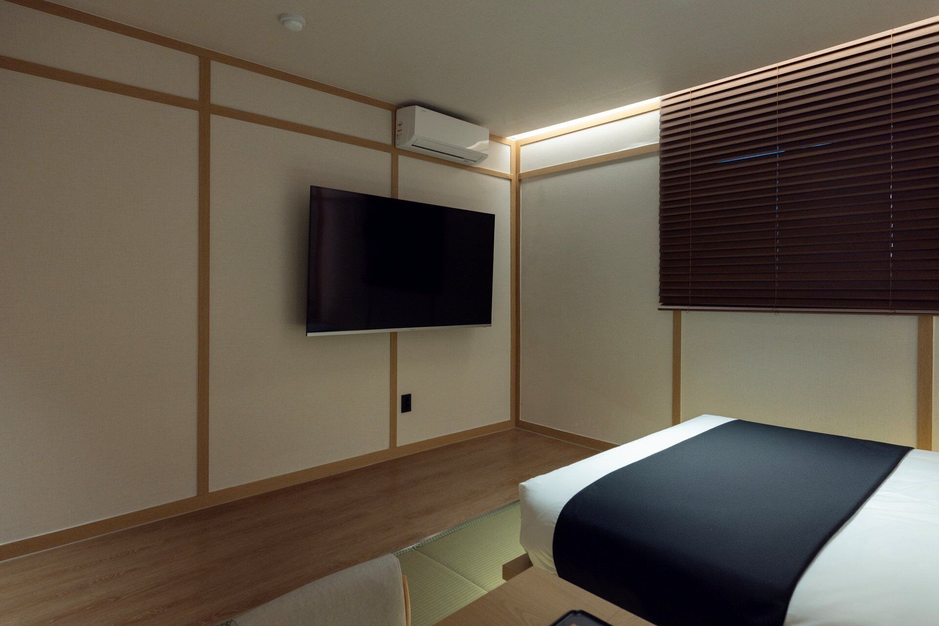 Habitación de hotel con cama, televisor de pantalla plana montado en la pared, paneles de madera y ventana con persianas.