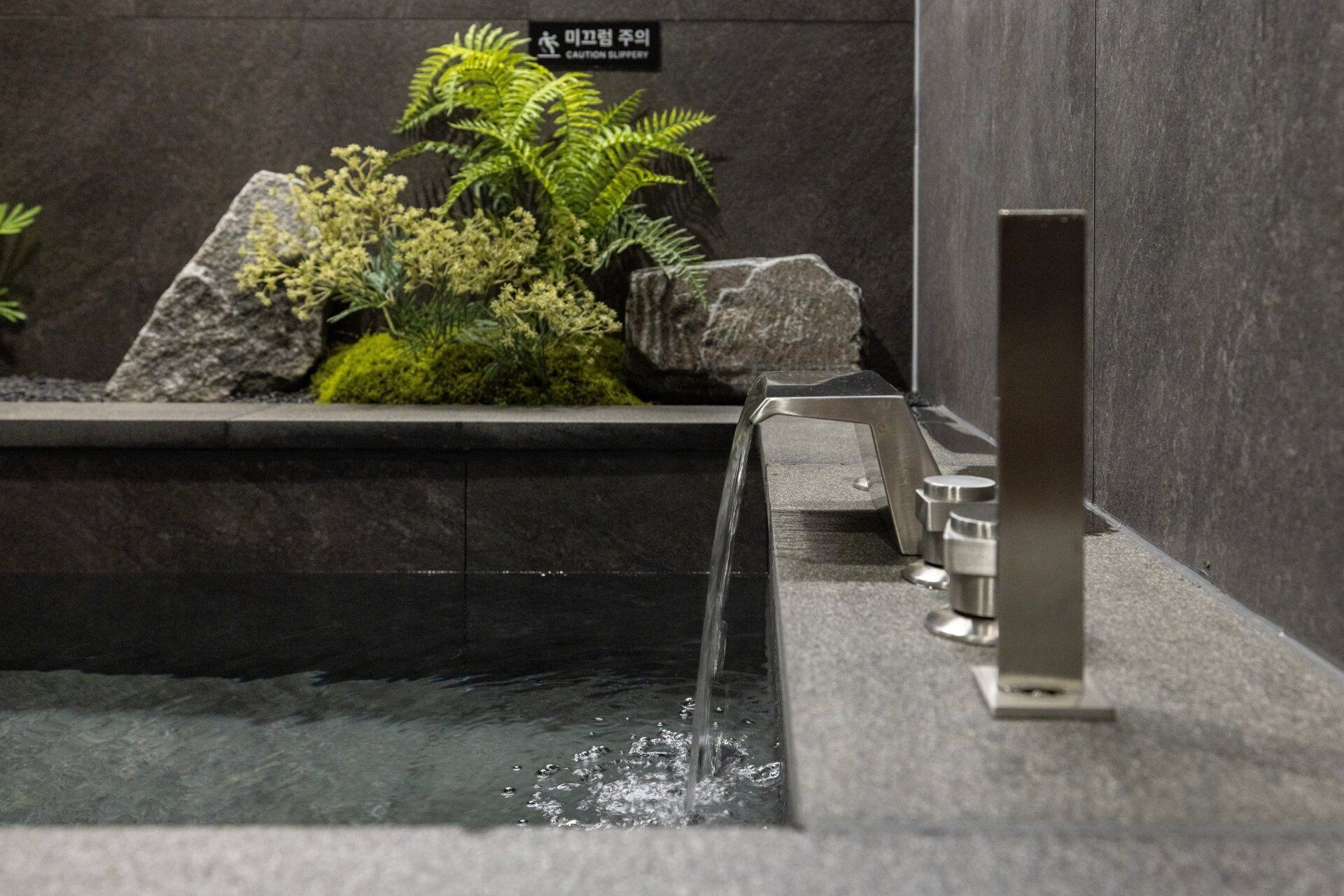 A water feature with a stream, a stone planter with plants, and a caution sign on the wall.