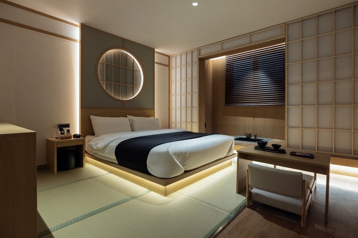A modern hotel room with a bed, bedside table, desk, chair, and a large window with blinds.