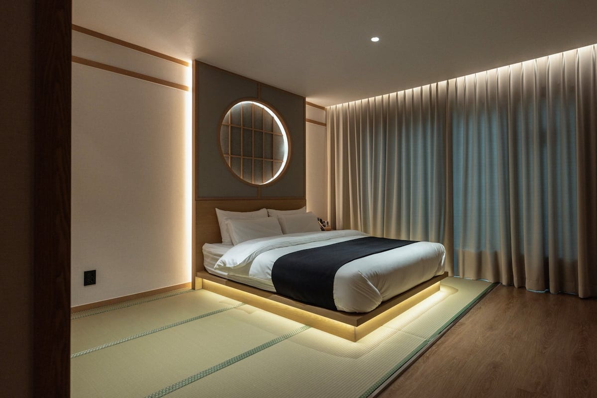 A modern hotel room with a bed, tatami mat flooring, a round wall-mounted light, and a large window with curtains.