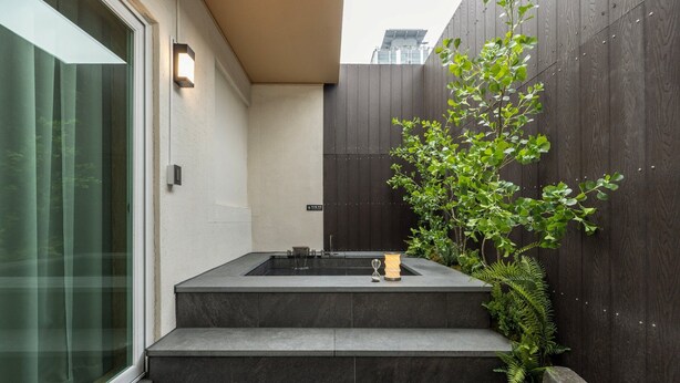 Un baño exterior moderno con un lavamanos de piedra, una pequeña plataforma elevada y una cerca de madera con follaje.