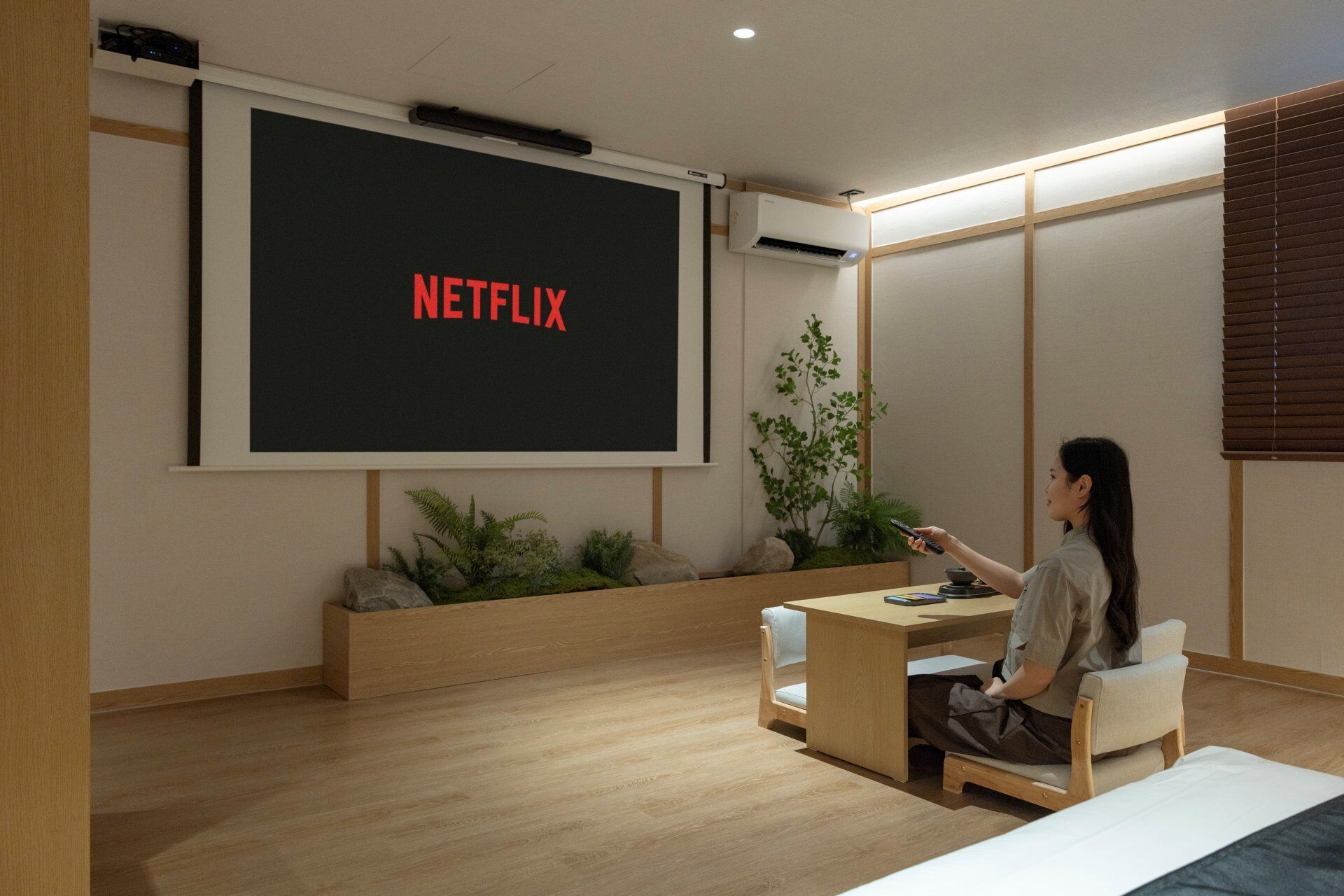 A room with a large screen displaying 'NETFLIX,' a wooden desk, a chair, and a small plant.
