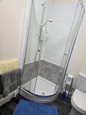 Ducha con cerramiento de vidrio, pared con azulejos blancos, piso gris, toalla amarilla y felpudo azul.