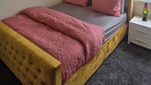 A neatly arranged bedroom with a tufted headboard, a bed with pink bedding, a decorative wall piece, and a bedside table with a lava lamp. - Glams 2beds Luxury Stylish Apartment (Wolverhampton)