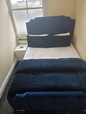 Una cama pequeña y ordenada con cabecera oscura y ropa de cama blanca en una habitación compacta con ventana.