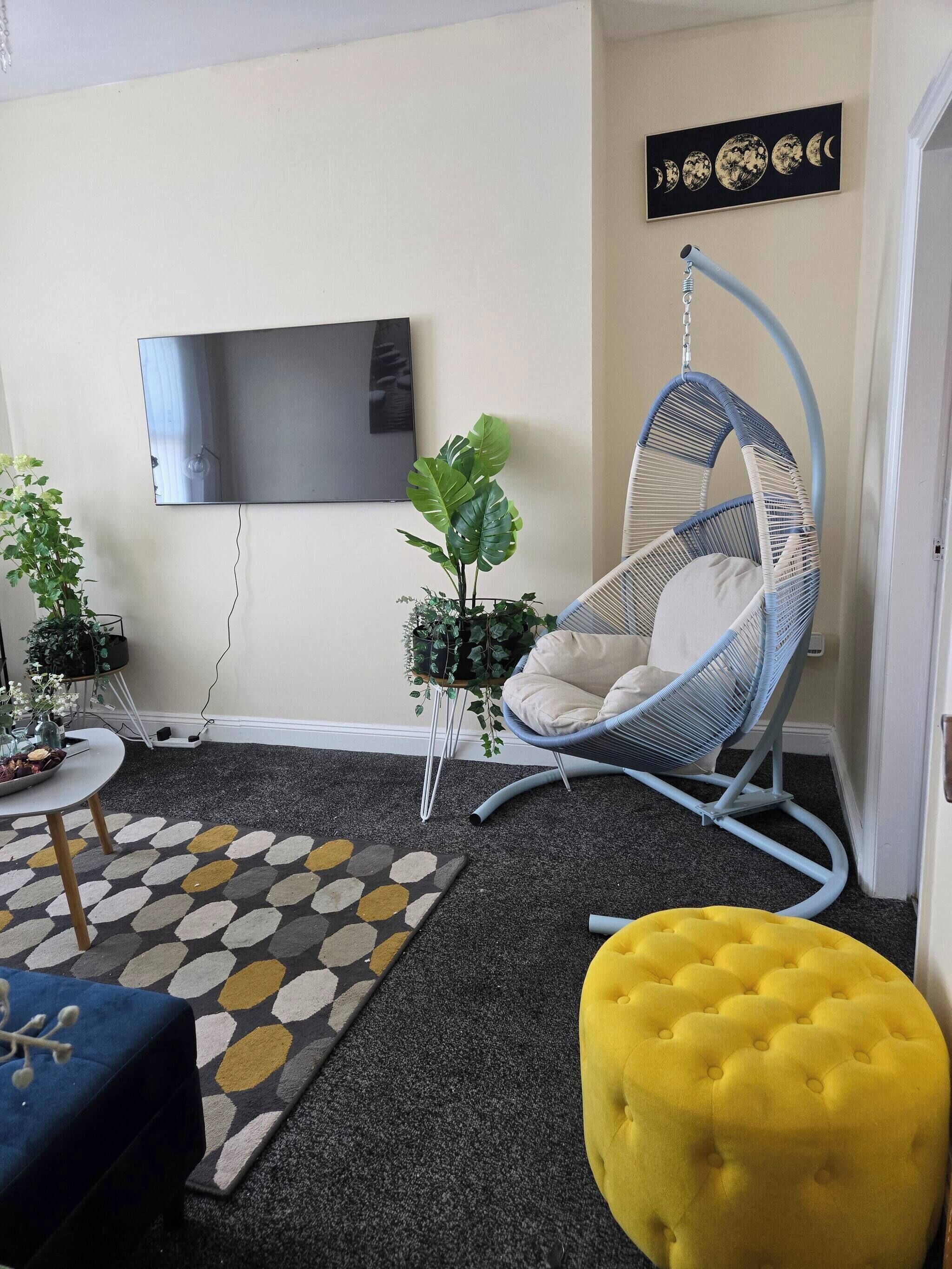 A living room with a hanging chair, a television mounted on the wall, a potted plant, and a yellow ottoman.