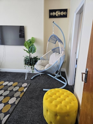 Een kamer met een blauwe hangstoel, een geel voetenbankje, een potplant en een aan de muur bevestigde televisie.