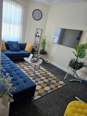 Un salon avec un canapé bleu, une table basse, un téléviseur et une horloge au mur.