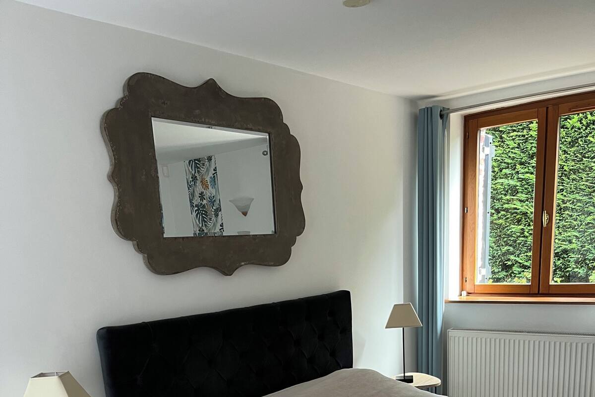 Un dormitorio con una cama grande, una mesita de noche con lámpara, un espejo y una ventana con cortinas.