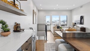 Una cucina moderna con un piano di lavoro bianco, mobili in legno e un forno integrato. C'è un'area pranzo con tavolo e sedie in legno, e un'ampia finestra che offre vista sulle montagne.