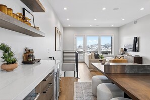 Eine moderne Küche mit weißer Arbeitsplatte, Holzschränken und einem eingebauten Ofen. Ein Essbereich mit Holztisch und -stühlen lädt ein, und ein großes Fenster bietet einen Blick auf die Berge.