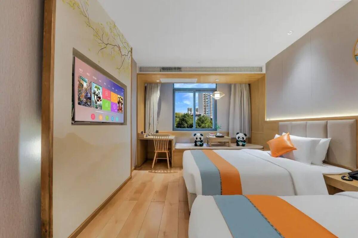 Habitación de hotel con dos camas, un escritorio, un ventanal amplio con vistas a edificios y un televisor con iconos coloridos.