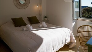 A bedroom with a large bed, two pillows, a bedside table with a lamp, a small plant, and a view of the outdoors through a window. - Azur Cancale (Cancale)