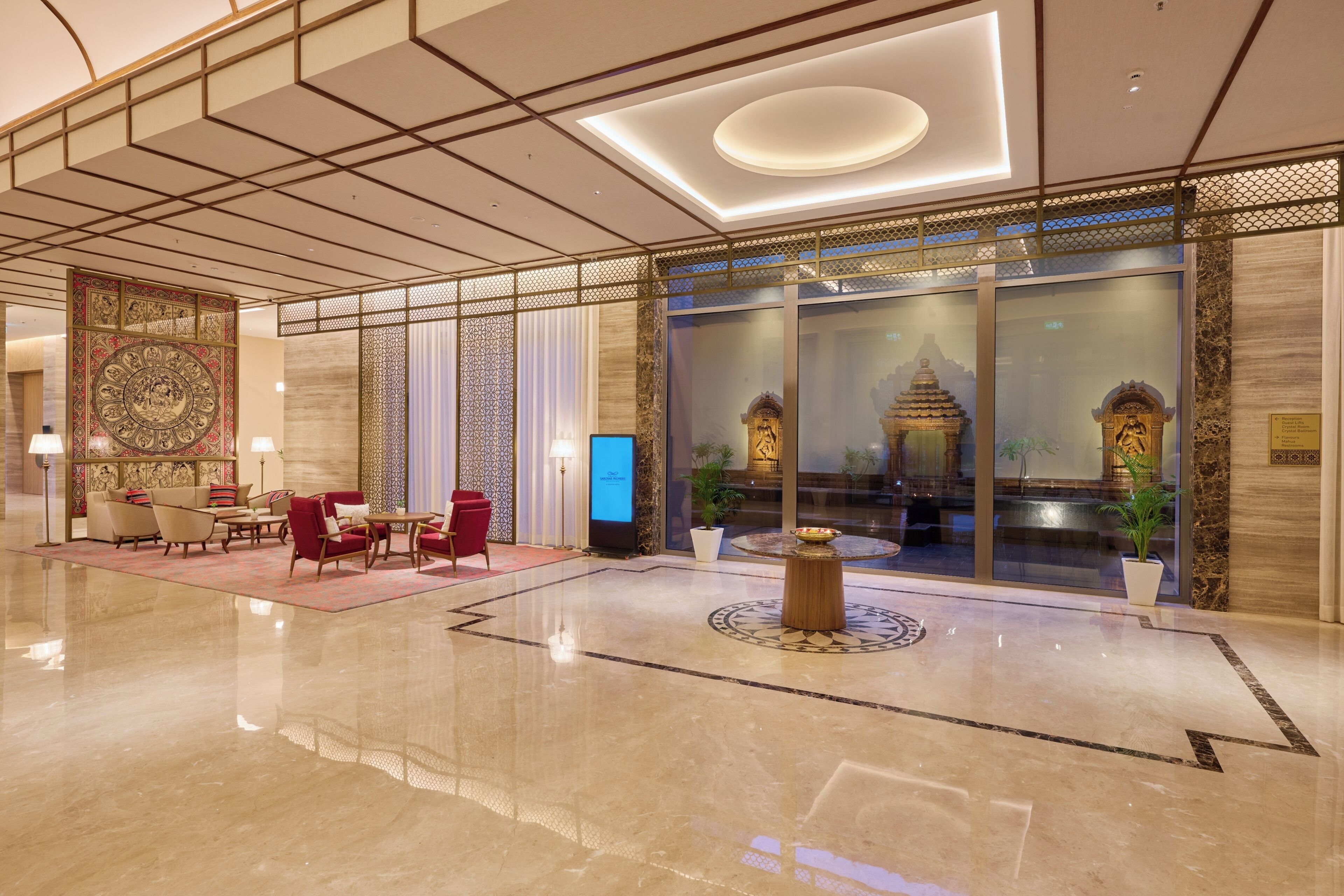 A lobby with a polished marble floor, a seating area with armchairs and a red sofa, a decorative wall with intricate patterns, and a large glass window showcasing a traditional building.