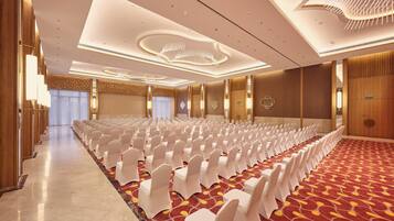 Un salón de conferencias con filas de sillas blancas, una alfombra estampada y un diseño moderno en el techo.