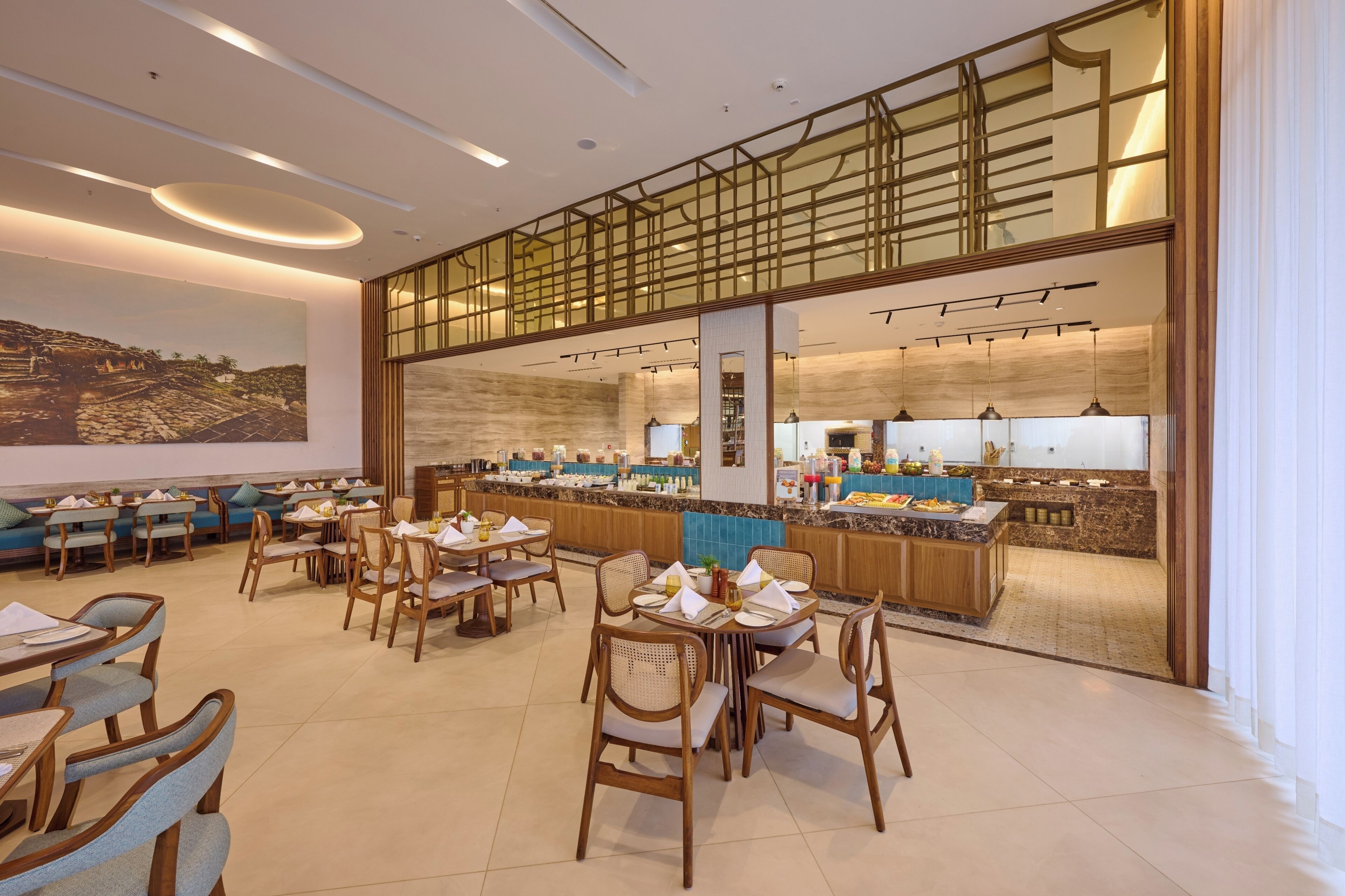 Un restaurant moderne doté de nombreuses tables et chaises, d’un espace cuisine visible à travers une paroi en verre et d’une grande fresque murale.