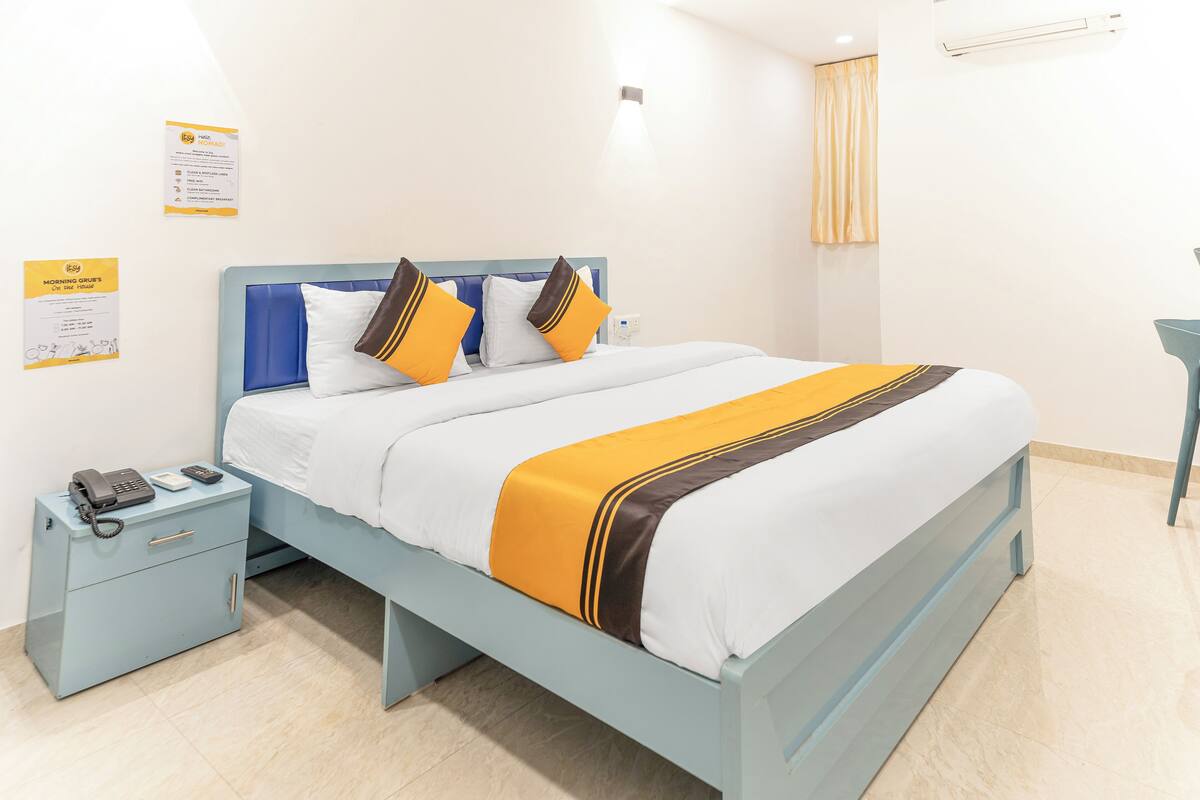 A neatly made bed with a blue headboard, a bedside table with a phone and a lamp, and a wall-mounted air conditioner.