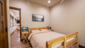 Une chambre avec un lit en bois, une table de chevet avec une lampe, un tableau au mur et une vue sur un couloir.