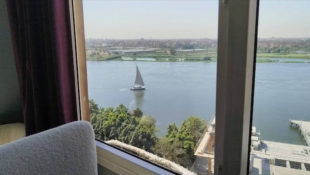 Une vue depuis une fenêtre donnant sur une rivière, avec un voilier, un paysage urbain en arrière-plan et une chaise au premier plan.