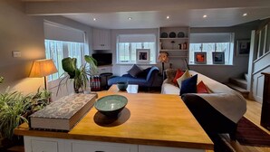 A modern living room with a wooden coffee table, a sofa, and a TV. There are plants, a lamp, and a bookshelf with books and decorative items. - The Smarden Snug for garden lovers (Smarden)