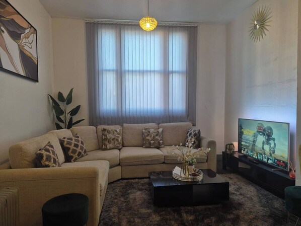 A living room with a beige sofa, a black coffee table, and a television displaying a program. - Glams one bedroom apartment (West Midlands)
