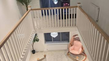 A staircase with white railings and a wooden handrail, leading to a landing with a large window, a chandelier, and a pink chair.