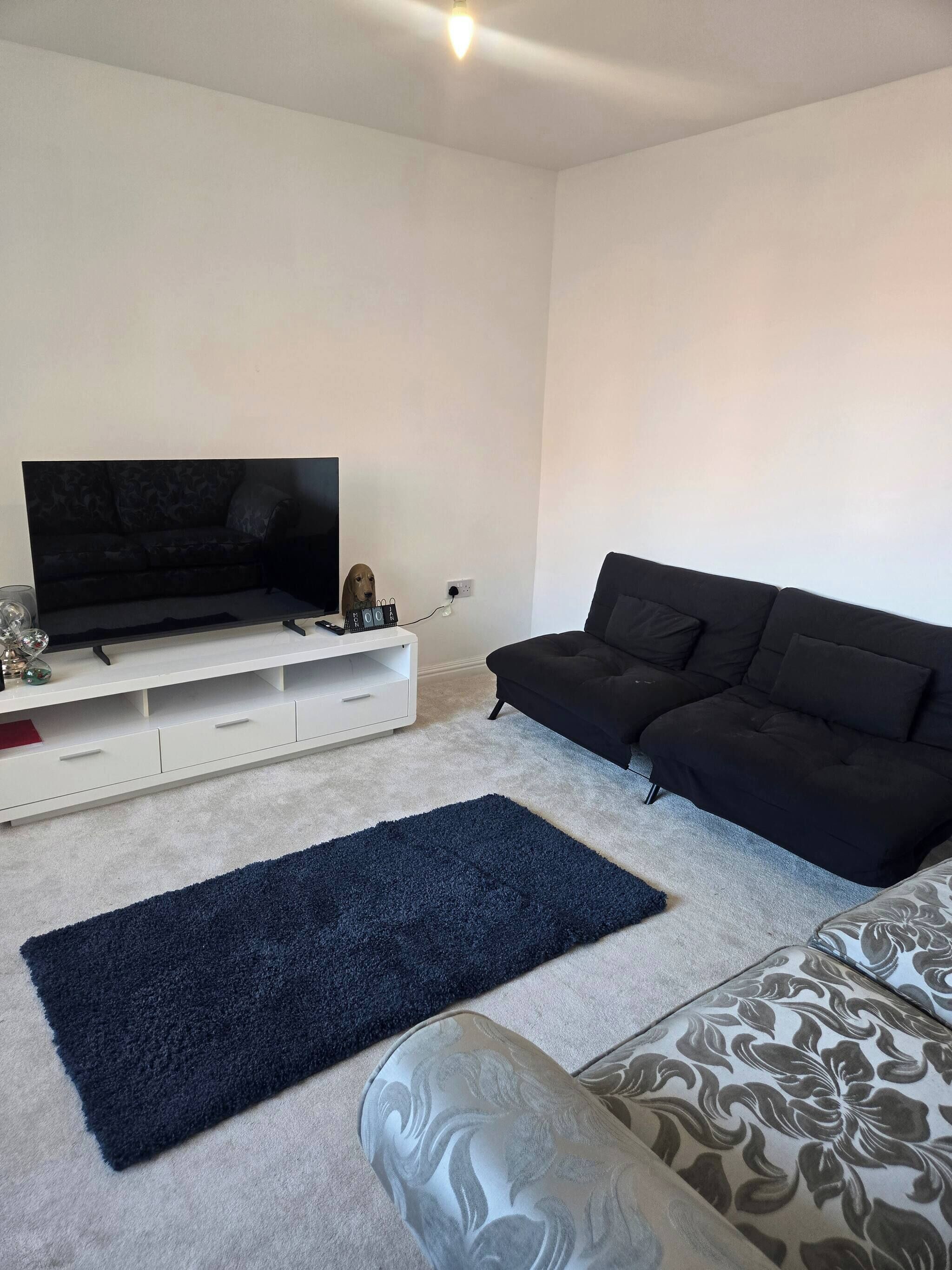 Un salon avec un téléviseur posé sur un meuble blanc, un canapé noir, un fauteuil à motifs et un tapis bleu foncé.