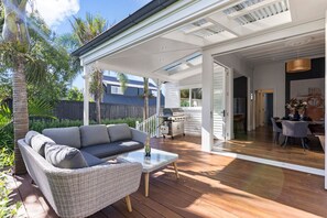 Eine überdachte Terrasse im Freien mit einer Rattan-Couch, einem Glastisch und einem Grill.