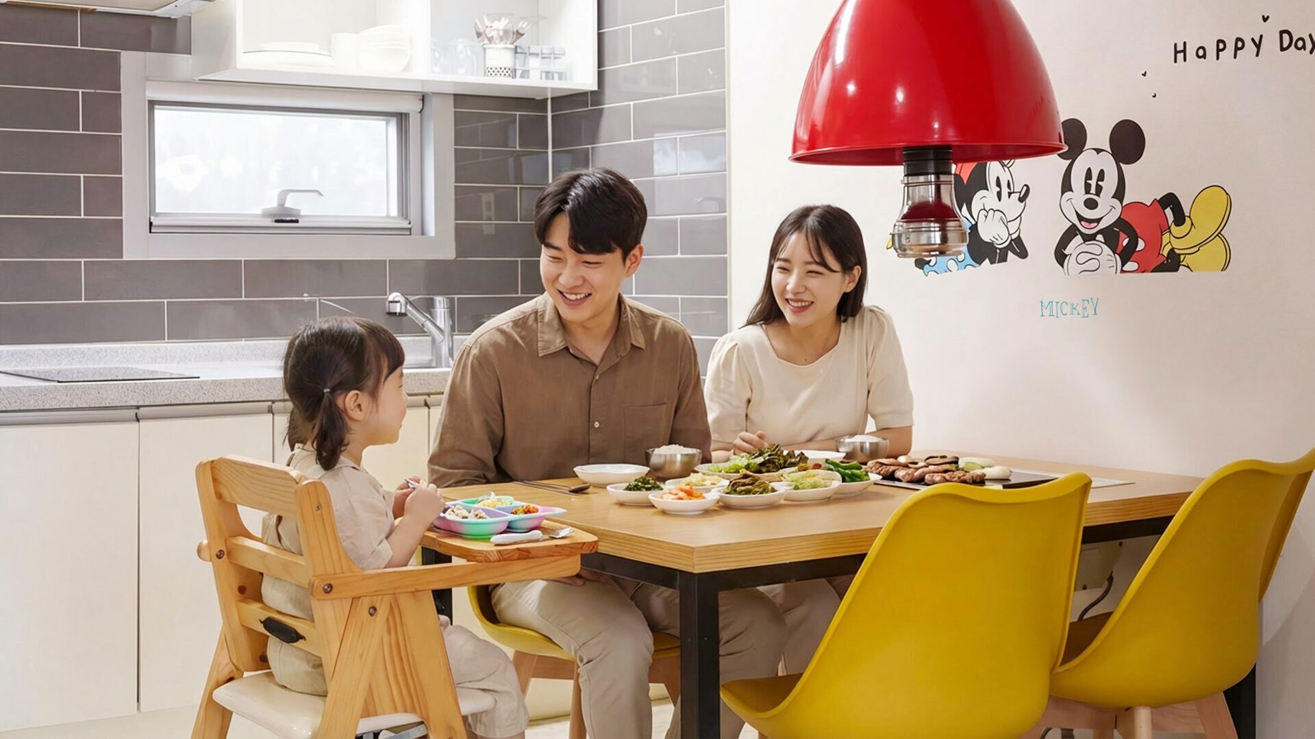 A family dining at a table with a Mickey Mouse mural, a red lamp, and a kitchen with grey tiles.