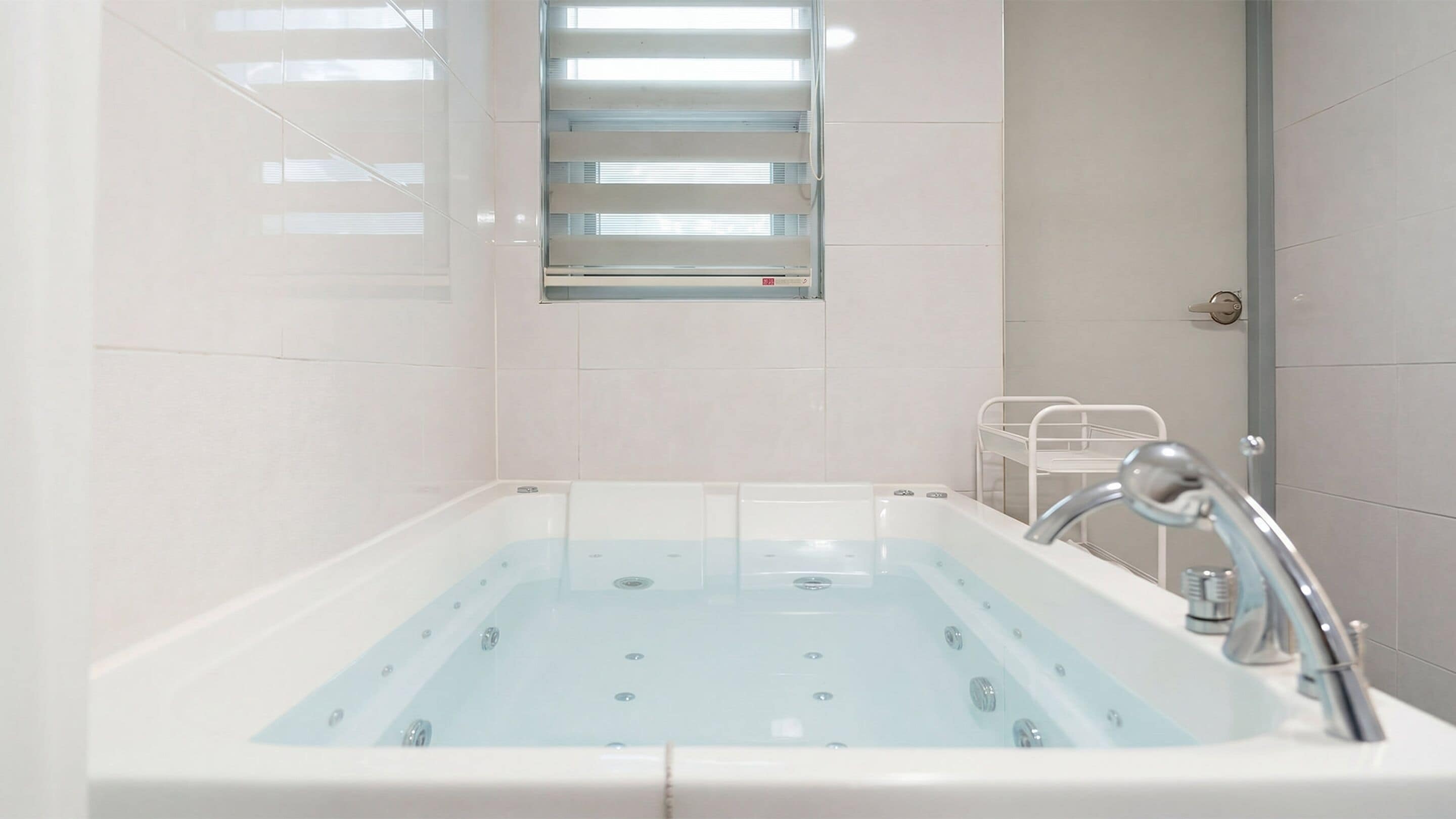 A modern bathroom with a large bathtub, a window with blinds, and a metal shelf unit.