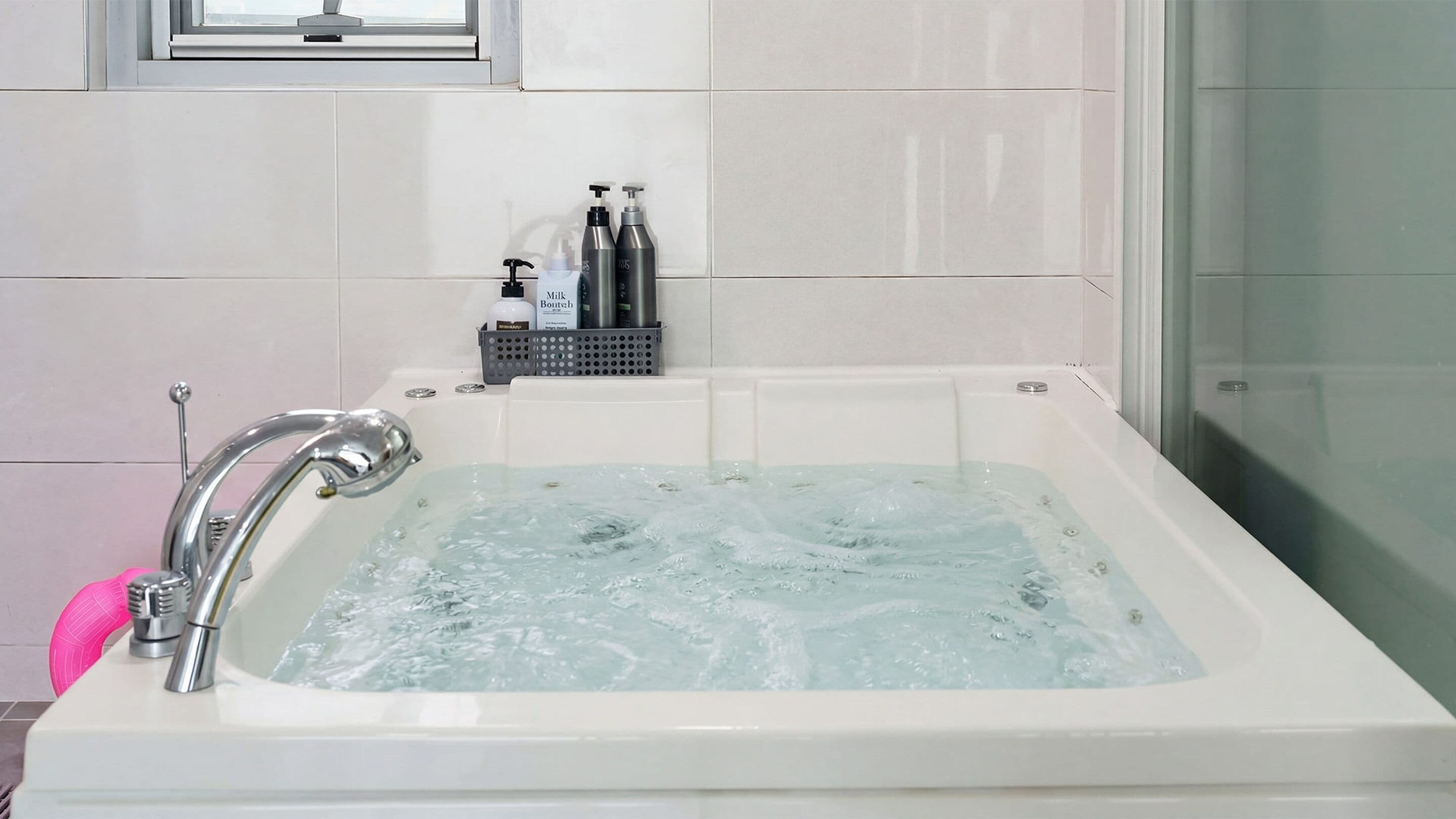 A modern bathroom with a bathtub filled with water, a shower area, and toiletries on a shelf.