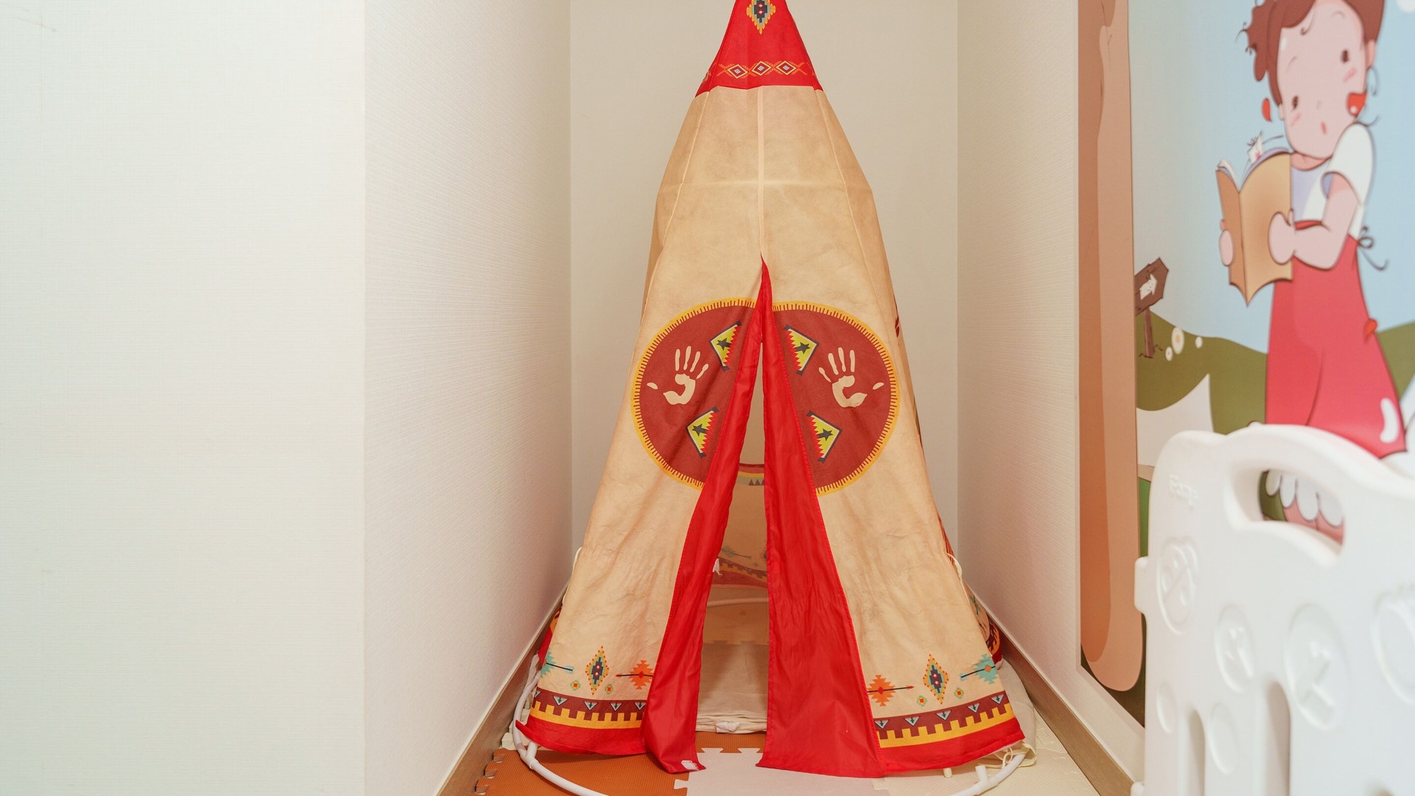 A tent-like structure with a red and beige design, possibly a play tent, in a room with a white wall and a cartoon character painting.
