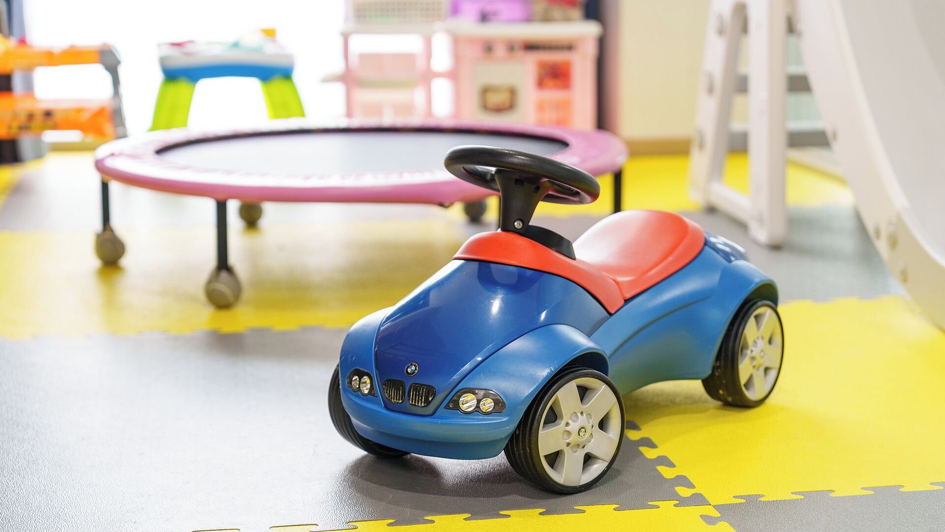 A toy car on a play mat with a trampoline and a small table in the background.