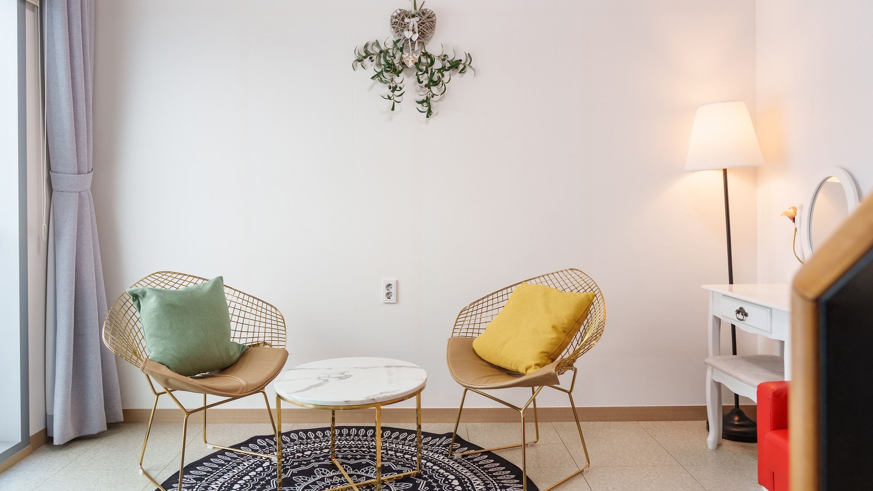 A room with two wireframe chairs with cushions, a small round table, a patterned rug, a wall-mounted light, and a white dresser with a mirror.