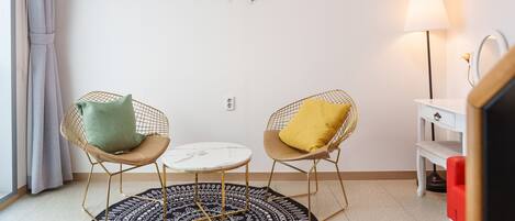 A room with two wireframe chairs with cushions, a small round table, a patterned rug, a wall-mounted light, and a white dresser with a mirror.