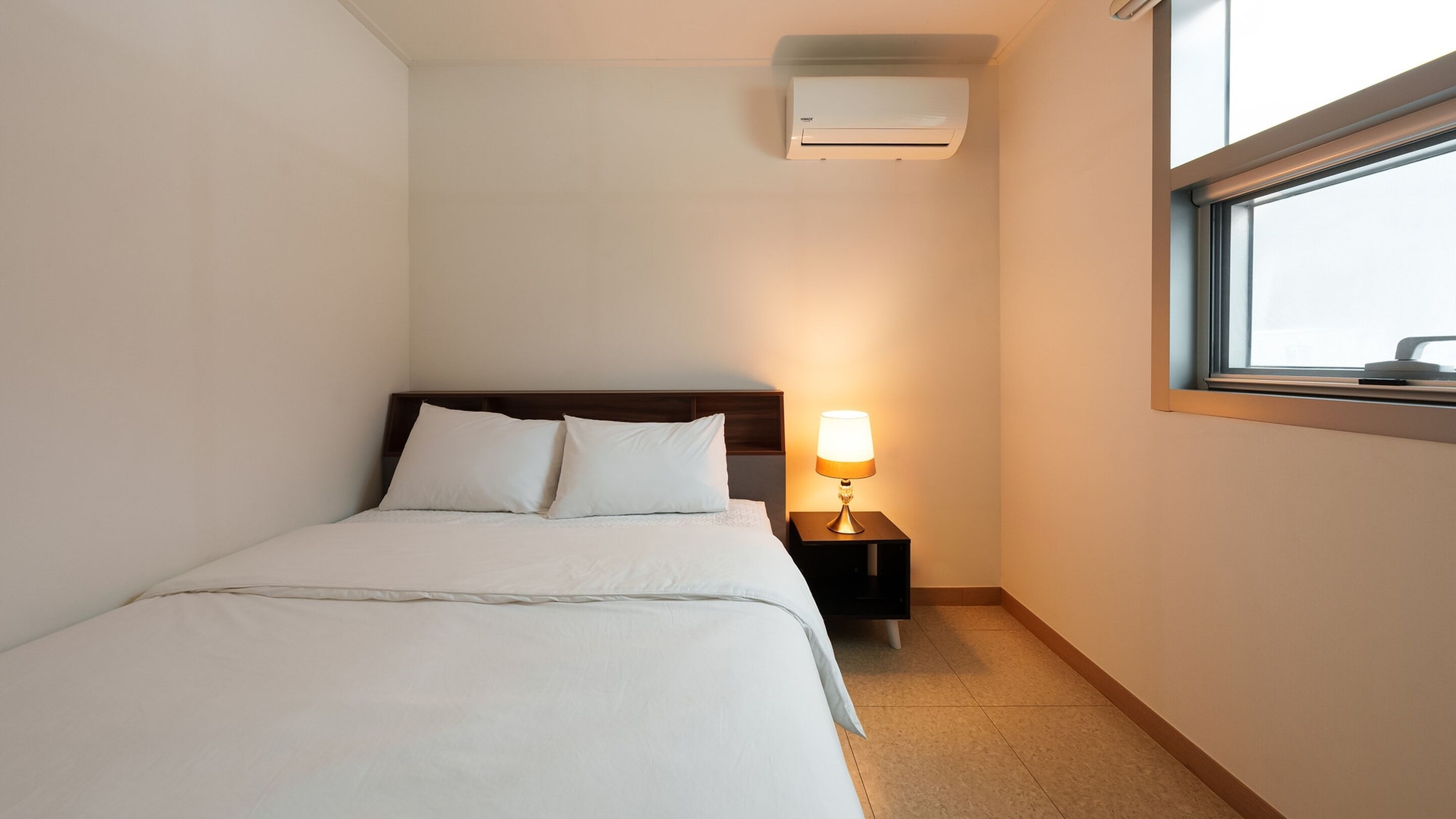 A hotel room with a bed, a nightstand with a lamp, an air conditioner, and a window.