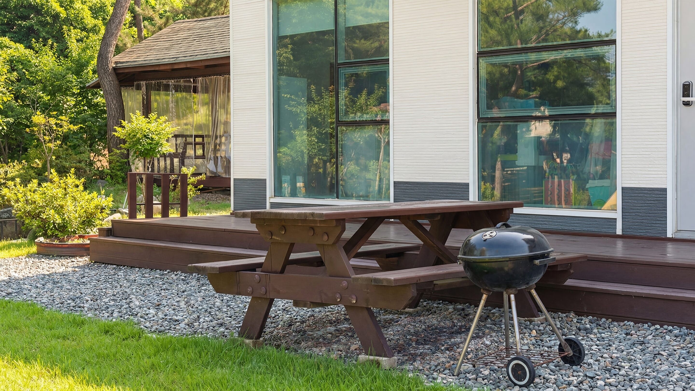 Una mesa de picnic de madera con una parrilla, ubicada sobre una zona de grava frente a una casa moderna con grandes ventanales.