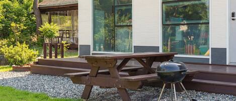 Una mesa de picnic de madera con una parrilla, ubicada sobre una zona de grava frente a una casa moderna con grandes ventanales.