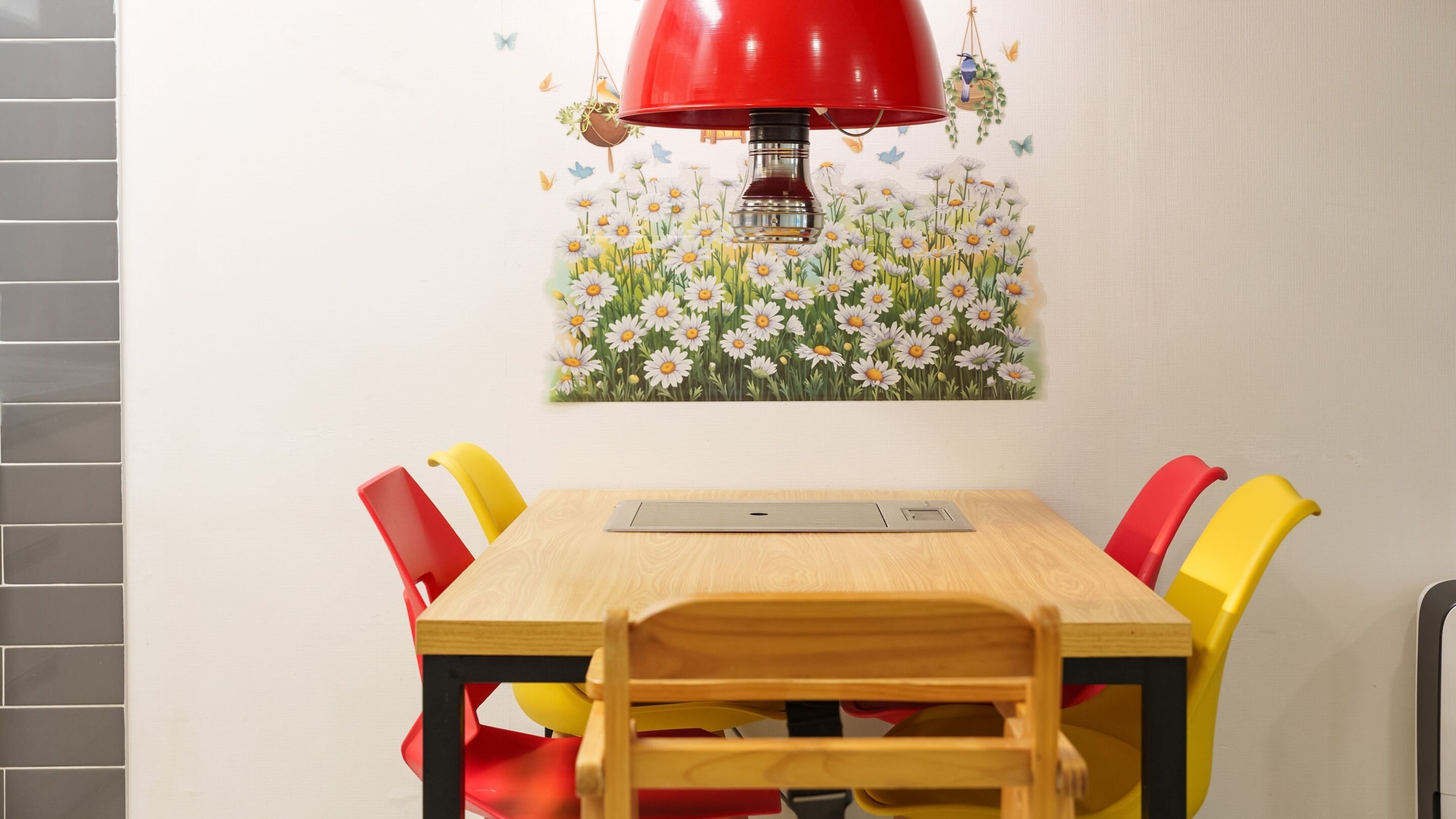 Un comedor con mesa de madera, sillas coloridas, una lámpara roja y un mural floral en la pared.