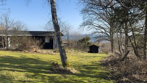 Une cabane rustique aux murs en bois, entourée d’un champ herbeux et d’arbres.