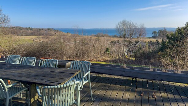 Une terrasse en bois avec une table et des chaises, offrant une vue sur un paysage côtier arboré et sur la mer.