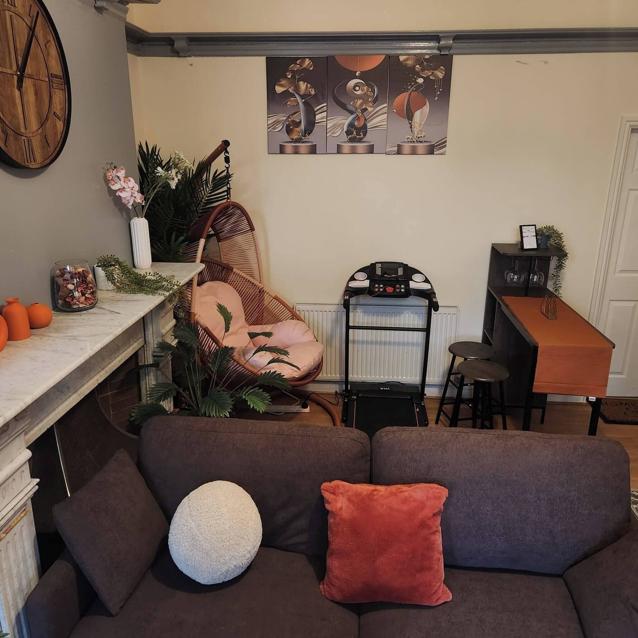 A cozy living area with a brown sofa, a white round pillow, and a red pillow. There's a wicker chair, a small table with a vase, and a wall clock.