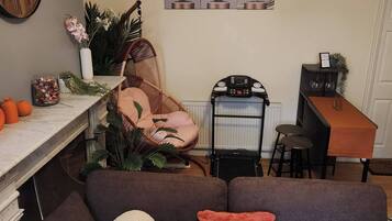 A cozy living area with a brown sofa, a white round pillow, and a red pillow. There's a wicker chair, a small table with a vase, and a wall clock.